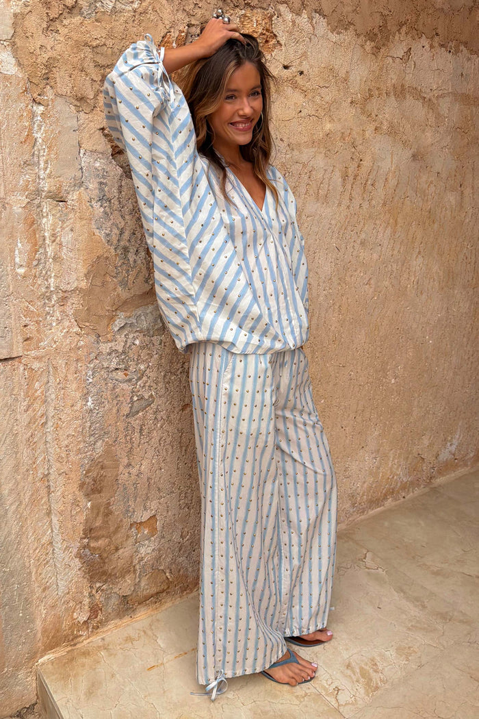 Blue and cream striped balloon trousers with gold studs and tie-cuffs, worn as a co-ord set against a stone wall.