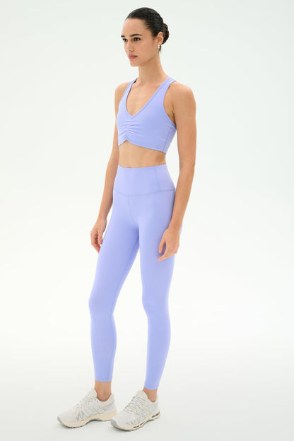A woman wears the SPLITS59 Airweight Bra with Ruching in Purple Haze and matching high-waisted leggings, paired with white athletic shoes, standing against a plain white background.
