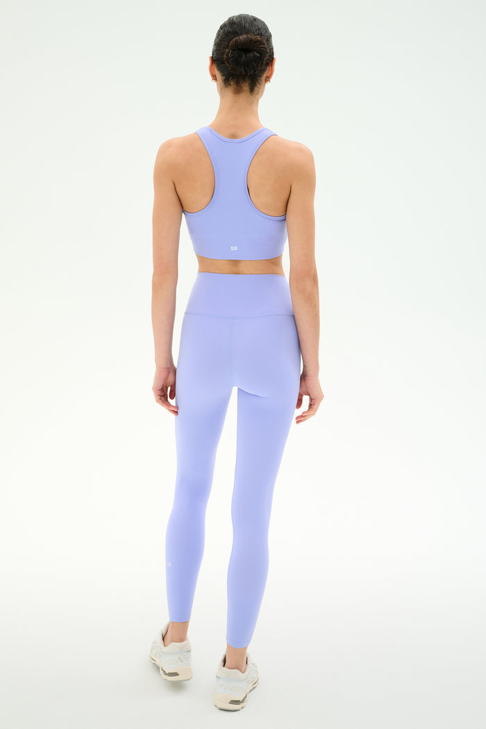 A person faces away, wearing the SPLITS59 Airweight Bra with Ruching in Purple Haze and matching high-waisted leggings with white sneakers, set against a plain light background.