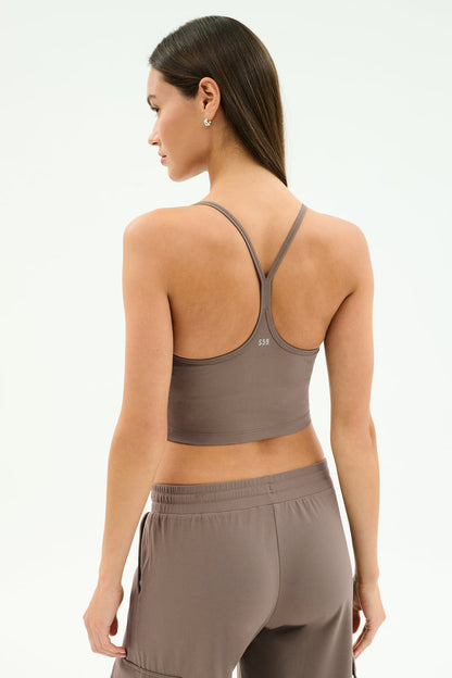 A woman with long brown hair stands against a plain light background wearing the SPLITS59 Airweight Crop in Lentil, paired with matching pants—perfect for hot yoga or any workout.