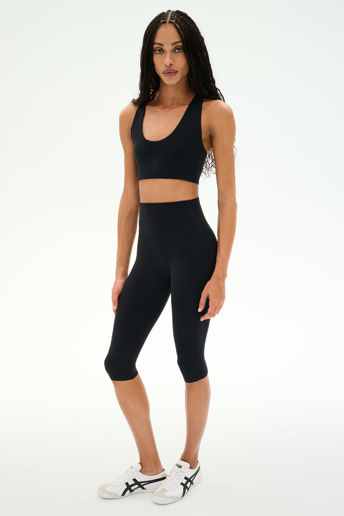 A person stands facing forward wearing a black sports bra, SPLITS59 Airweight High Waist 16" Capri Legging in black, and white sneakers with black accents against a plain white background.