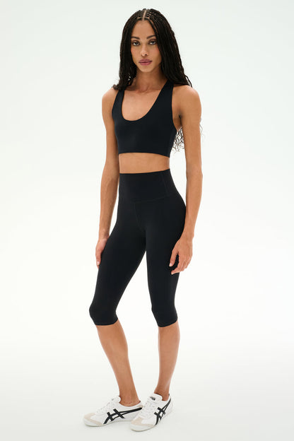 A person stands facing forward wearing a black sports bra, SPLITS59 Airweight High Waist 16" Capri Legging in black, and white sneakers with black accents against a plain white background.