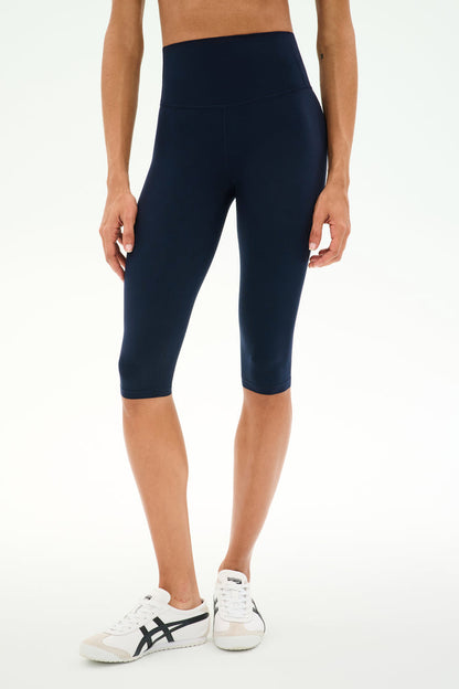 A person wearing SPLITS59 Airweight High Waist 16" Capri Legging in Indigo and white sneakers, shown from shoulders to feet, standing against a plain white background.