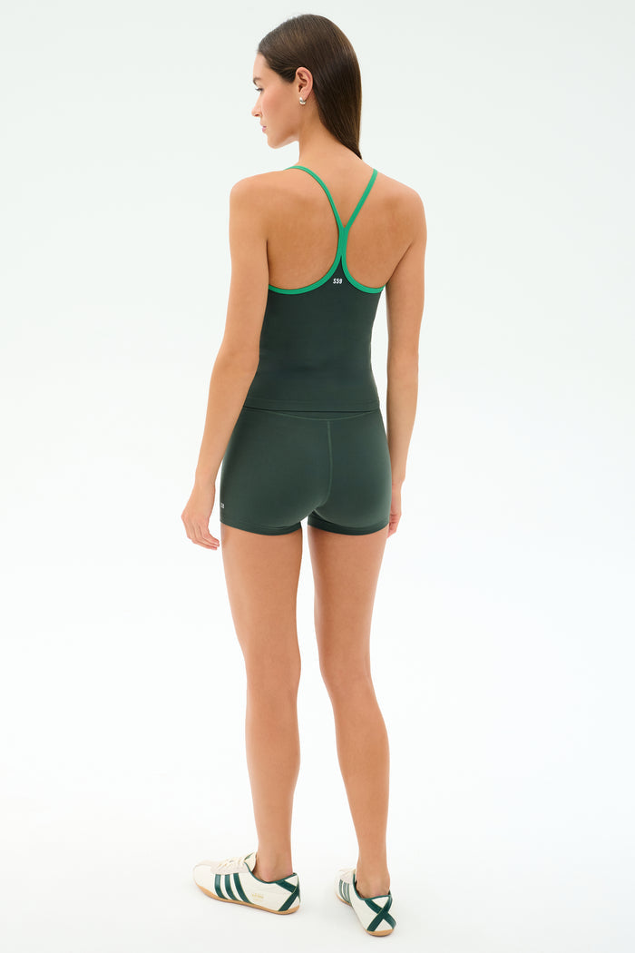 A woman faces away, wearing SPLITS59’s Airweight High Waist 3.5” Short in Military and a matching dark green tank top with green trim, paired with white sneakers with green stripes, set against a plain light background.