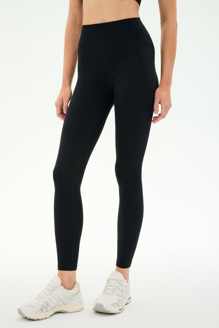 A person wearing SPLITS59 Airweight High Waist Legging in black and white sneakers stands against a plain light background—perfect for hot yoga or all-day comfort thanks to the soft Airweight fabric.