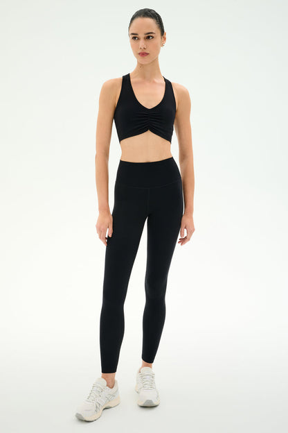 A person stands against a plain background wearing the SPLITS59 Airweight High Waist Legging - Black, perfect for hot yoga, paired with a black sports bra and white sneakers.