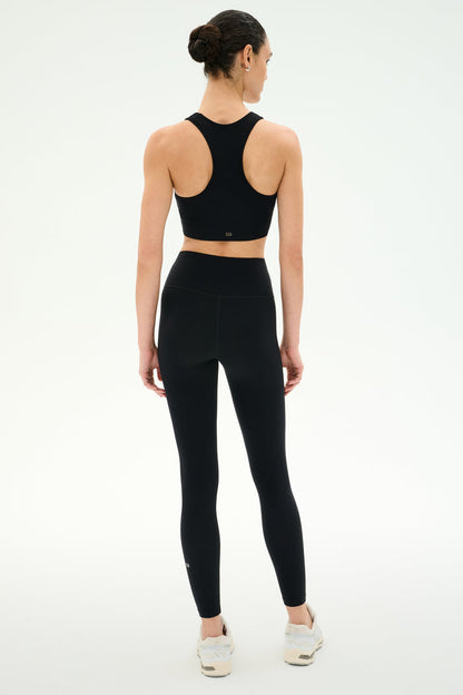 A woman faces away wearing SPLITS59's Airweight High Waist Legging in black with a matching sports bra and white sneakers against a plain, light background—ideal for hot yoga sessions.