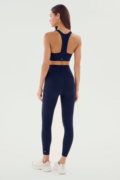 A woman stands with her back to the camera wearing the SPLITS59 Airweight High Waist Legging in Indigo and a matching navy blue sports bra, complemented by white sneakers, against a plain background.