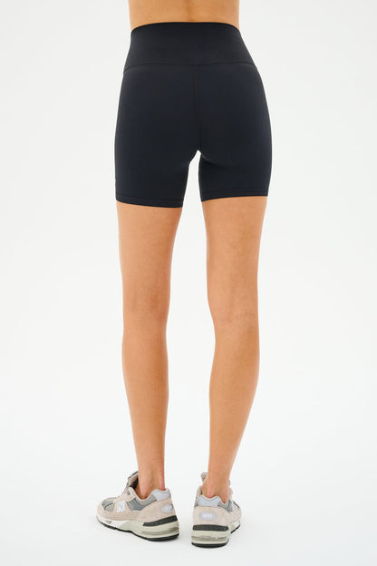 Viewed from the back, a person wears SPLITS59's Airweight High Waist Short in black and gray sneakers, ready for cycling against a plain white backdrop.