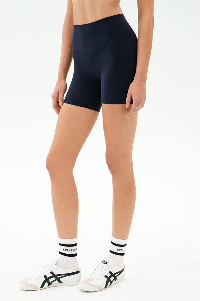 A person wears SPLITS59's Airweight High Waist Short in Indigo, a seamless tank workout top, white socks, and white sneakers with black stripes while standing against a plain white background.