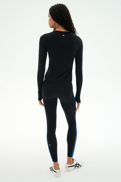 A person stands facing away, wearing the SPLITS59 Airweight Lite Long Sleeve Waist Length top in black and matching leggings with blue side stripes, paired with white sneakers against a plain light background—an ideal outfit for gym workouts.