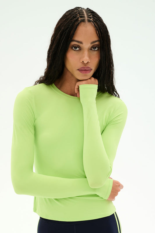 A woman with long braided hair wears the SPLITS59 Airweight Lite Long Sleeve Waist Length tee in Limeade, standing against a white background while resting her chin on her right hand and looking at the camera. The lightweight, quick-dry fabric makes this top ideal for gym workouts or everyday wear.