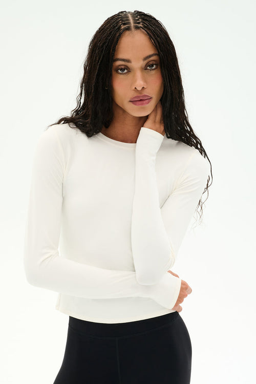 A person with long braided hair models the SPLITS59 Airweight Lite Long Sleeve Waist Length tee in white, paired with black pants, posing with one hand touching their face against a plain white background.