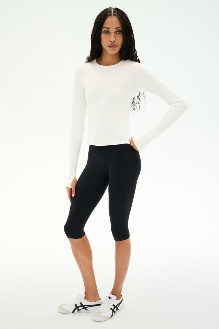 A woman wears the SPLITS59 Airweight Lite Long Sleeve Waist Length tee in white, paired with black knee-length leggings and white sneakers, standing against a plain white background—an ideal outfit for gym workouts.