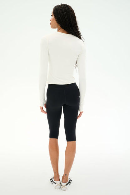 A woman stands facing away from the camera, wearing the SPLITS59 Airweight Lite Long Sleeve Waist Length top in white, paired with black knee-length leggings ideal for gym workouts and white sneakers, against a plain white background.