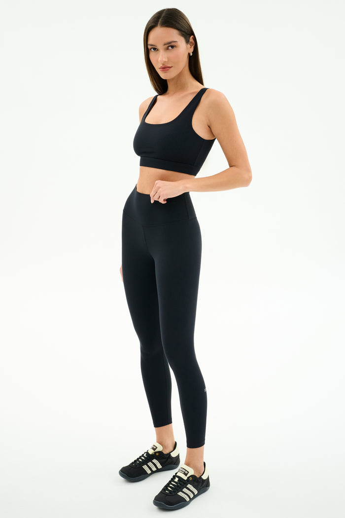 A woman stands against a white background wearing the SPLITS59 Airweight Rib Bra in black, paired with high-waisted leggings and black Adidas sneakers—an ideal outfit for hot yoga sessions.