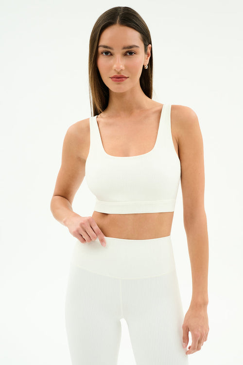 A woman wears the SPLITS59 Airweight Rib Bra in white, crafted from moisture-wicking ribbed fabric—ideal for hot yoga—paired with matching high-waisted leggings, standing against a plain white background.