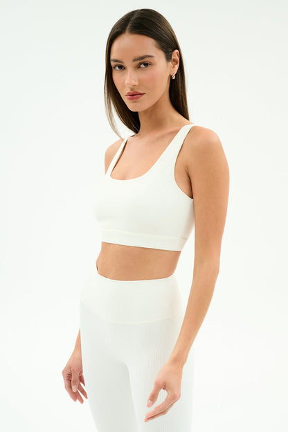A woman wears the SPLITS59 Airweight Rib Bra in white, standing against a plain background—this ribbed, moisture-wicking bra is perfect for hot yoga thanks to its comfortable design.