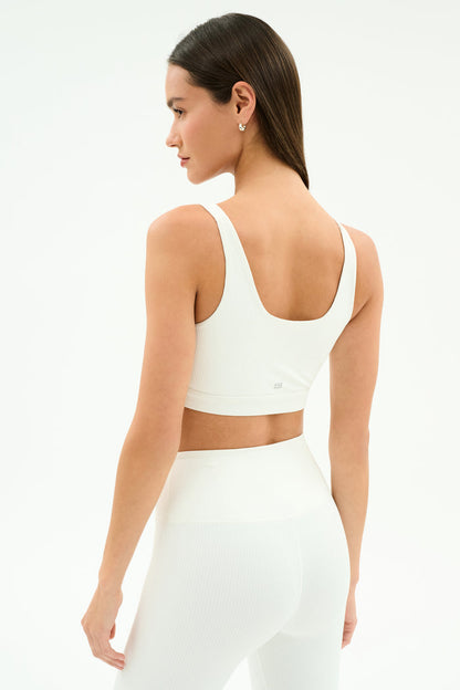 A woman models the SPLITS59 Airweight Rib Bra in white, turning to her left to showcase its breathable ribbed fabric—ideal for hot yoga—against a plain white background.