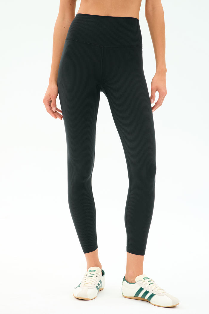 A person wears SPLITS59 Airweight Rib High Waist 7/8 leggings in black—ideal for Pilates or hot yoga—with white sneakers featuring green stripes, standing against a simple light background.