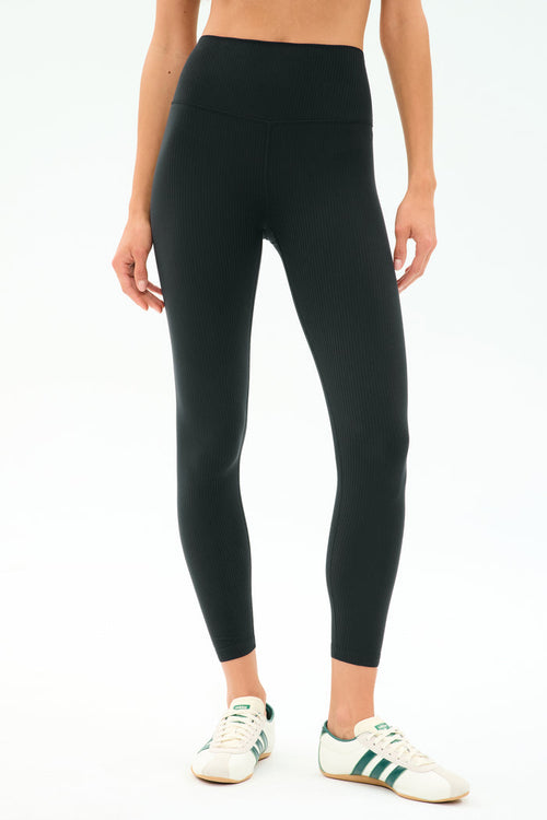 A person wears SPLITS59 Airweight Rib High Waist 7/8 leggings in black—ideal for Pilates or hot yoga—with white sneakers featuring green stripes, standing against a simple light background.
