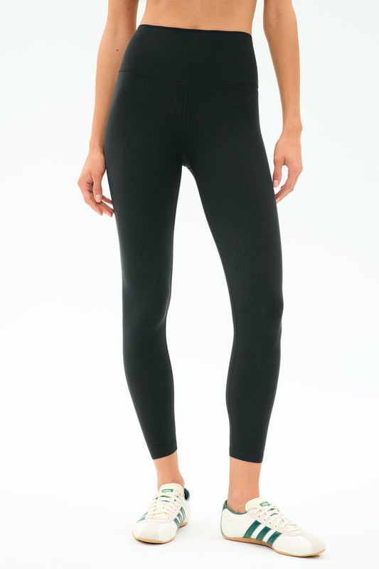 A person wears SPLITS59 Airweight Rib High Waist 7/8 leggings in black—ideal for Pilates or hot yoga—with white sneakers featuring green stripes, standing against a simple light background.