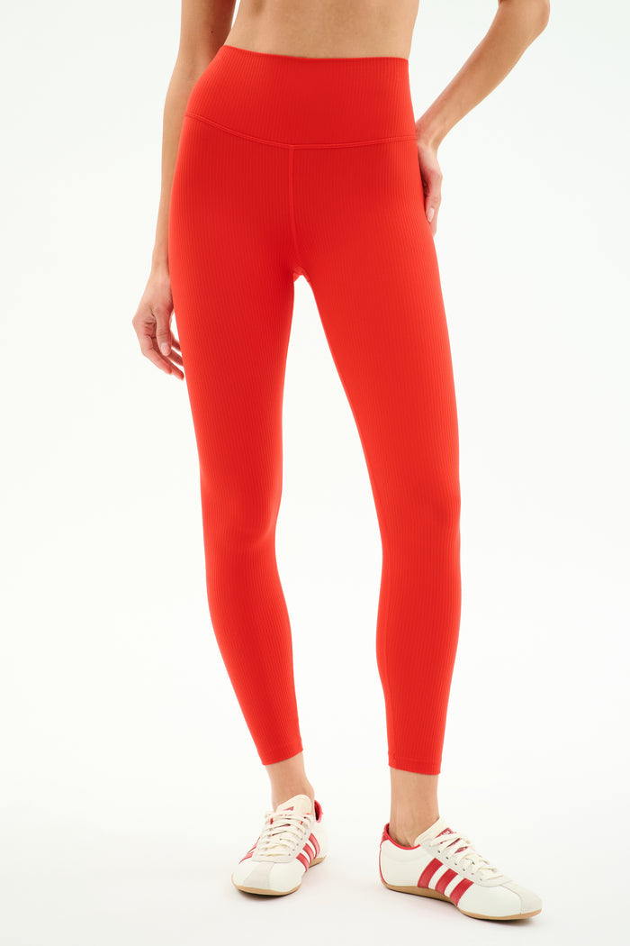 A person wearing SPLITS59 Airweight Rib High Waist 7/8 leggings in Lotus Red with red and white sneakers stands against a plain white background.
