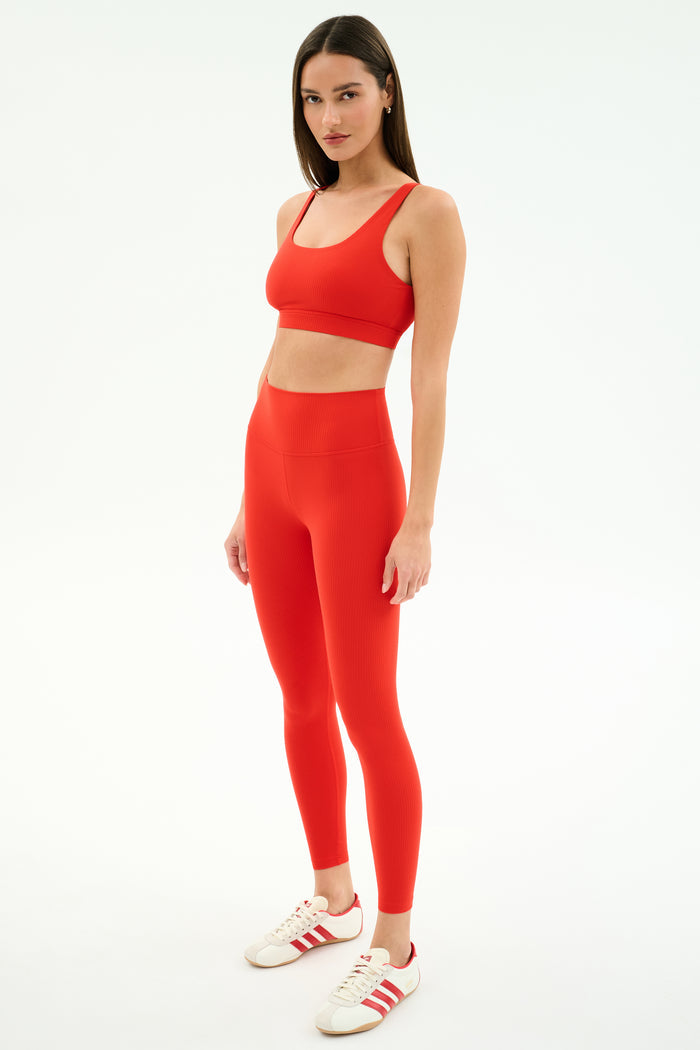 A woman stands against a white background wearing SPLITS59 Airweight Rib High Waist 7/8 leggings in Lotus Red, paired with a matching red sports bra and white-red sneakers.