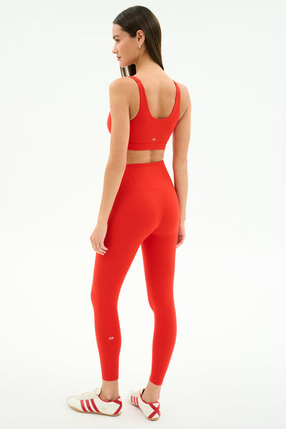 A woman, facing away and looking to the side, wears SPLITS59 Airweight Rib High Waist 7/8 leggings in Lotus Red with a matching sports bra and white Adidas sneakers against a plain white background.