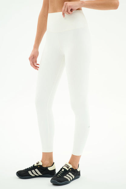 A person wearing SPLITS59 Airweight Rib High Waist 7/8 leggings in white and black Adidas sneakers stands against a plain white background.