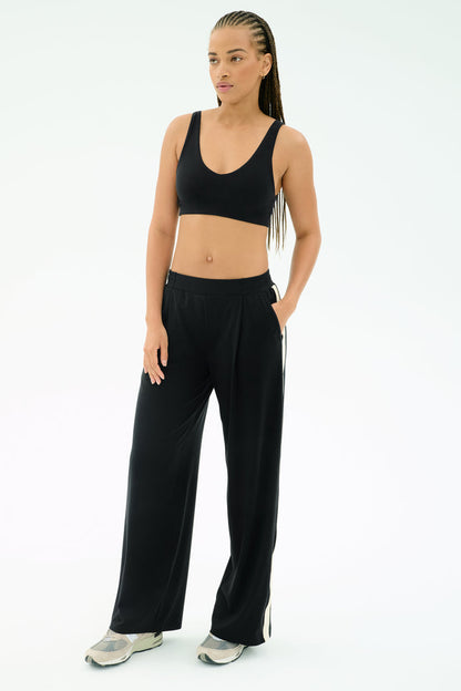 A woman poses against a plain background wearing the SPLITS59 Airweight Scoop Back Bra in black, paired with black wide-leg pants and sneakers, one hand in her pocket.