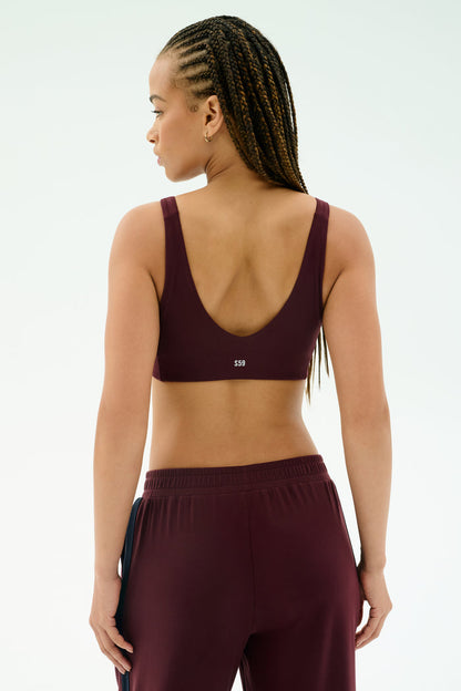 A person with braided hair is seen from the back wearing the SPLITS59 Airweight Scoop Back Bra in Dark Cherry, crafted from nylon spandex, against a light background.