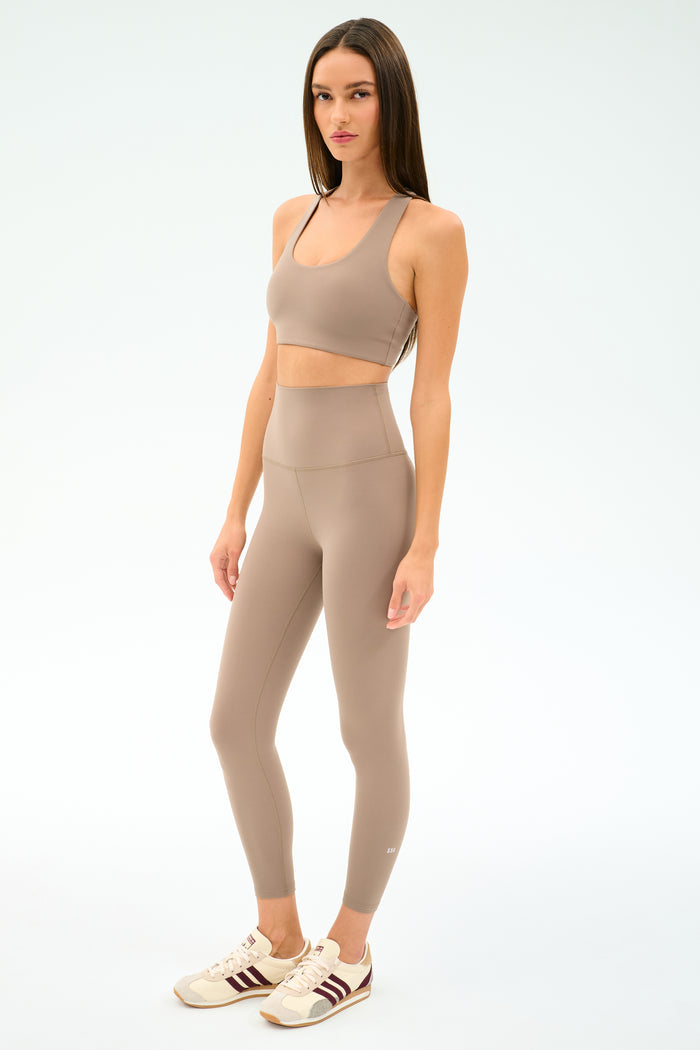 A woman poses against a white background wearing the SPLITS59 Airweight Super High Waist 7/8 leggings in Latte, paired with a taupe sports bra and beige sneakers featuring maroon stripes—an ideal outfit for hot yoga.