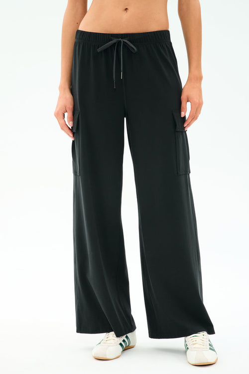 A person wearing SPLITS59’s Alex Airweight Cargo Pant in black, featuring side pockets and soft Airweight fabric, paired with white sneakers and standing against a plain light background.
