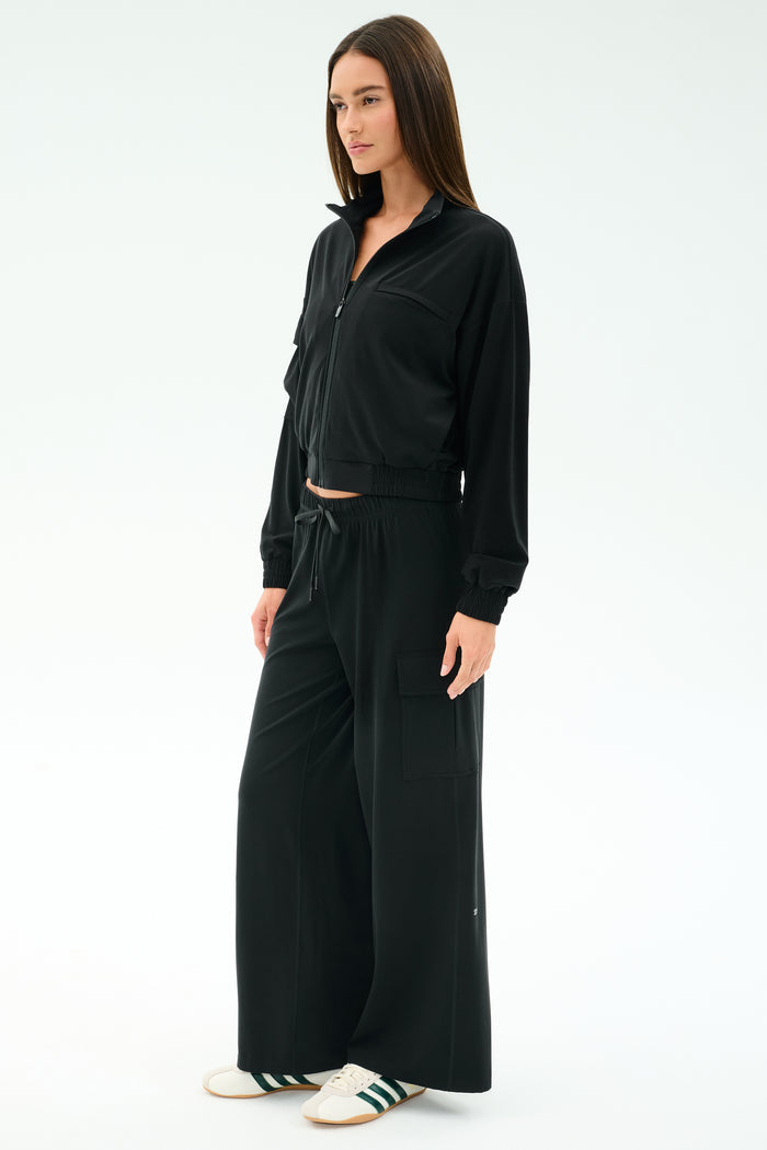 A woman stands against a plain white background wearing SPLITS59's black Alex Airweight Cargo Pant, styled with a matching black zip-up jacket and white sneakers featuring green stripes.