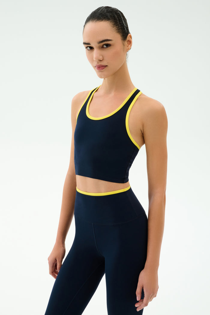 A woman poses against a plain backdrop in the SPLITS59 Amber Airweight Bralette in Indigo/Ray, featuring a scoop neck and yellow trim. Made from Airweight performance fabric, it's ideal for hot yoga sessions.