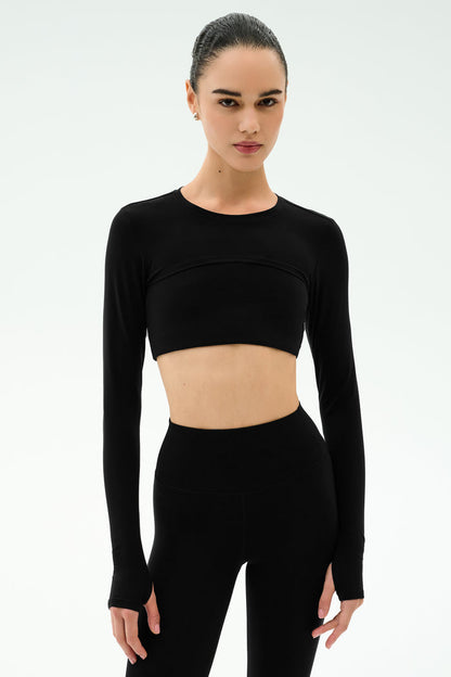 A woman stands against a plain white background wearing the SPLITS59 Amelia Airweight Lite Shrug in black with matching high-waisted leggings.