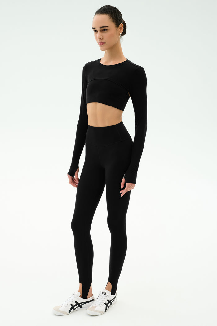 A person stands against a white background wearing the SPLITS59 Amelia Airweight Lite Shrug in black, paired with black long-sleeve crop top, black high-waisted stirrup leggings, and white athletic shoes.