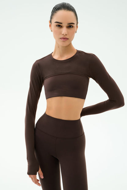A person wears the SPLITS59 Amelia Airweight Lite Shrug in Dark Chocolate, styled with high-waisted leggings, perfect for yoga or dance, against a plain light background.
