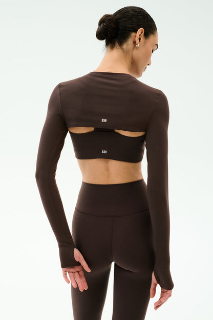 A woman stands with her back to the camera in SPLITS59’s Amelia Airweight Lite Shrug in Dark Chocolate, featuring long sleeves and back cutouts, paired with matching high-waisted leggings—ideal for yoga or dance.