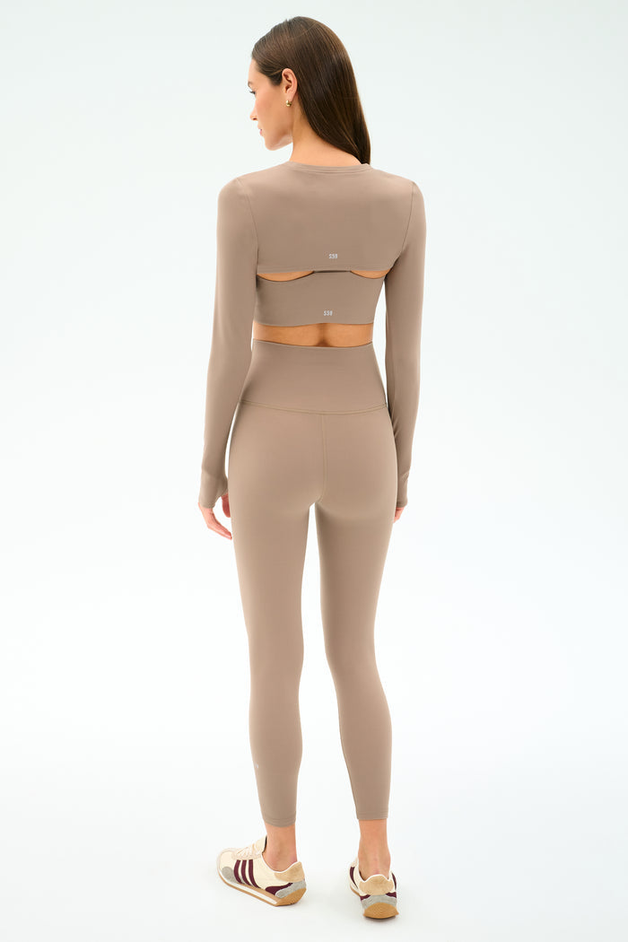 A woman faces away, wearing the SPLITS59 Amelia Airweight Lite Shrug in Latte—ideal for dance warmups or yoga—with white and maroon sneakers against a plain light background.