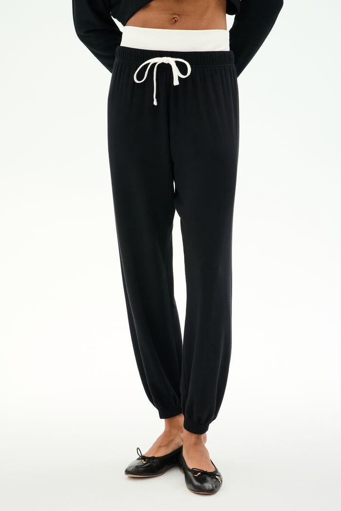 A person stands against a plain light background wearing the SPLITS59 Andie Dual Waist Sweatpant in Black/White—crafted from a soft modal blend with a white drawstring waistband—paired with black ballet flats.