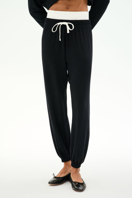 A person stands against a plain light background wearing the SPLITS59 Andie Dual Waist Sweatpant in Black/White—crafted from a soft modal blend with a white drawstring waistband—paired with black ballet flats.