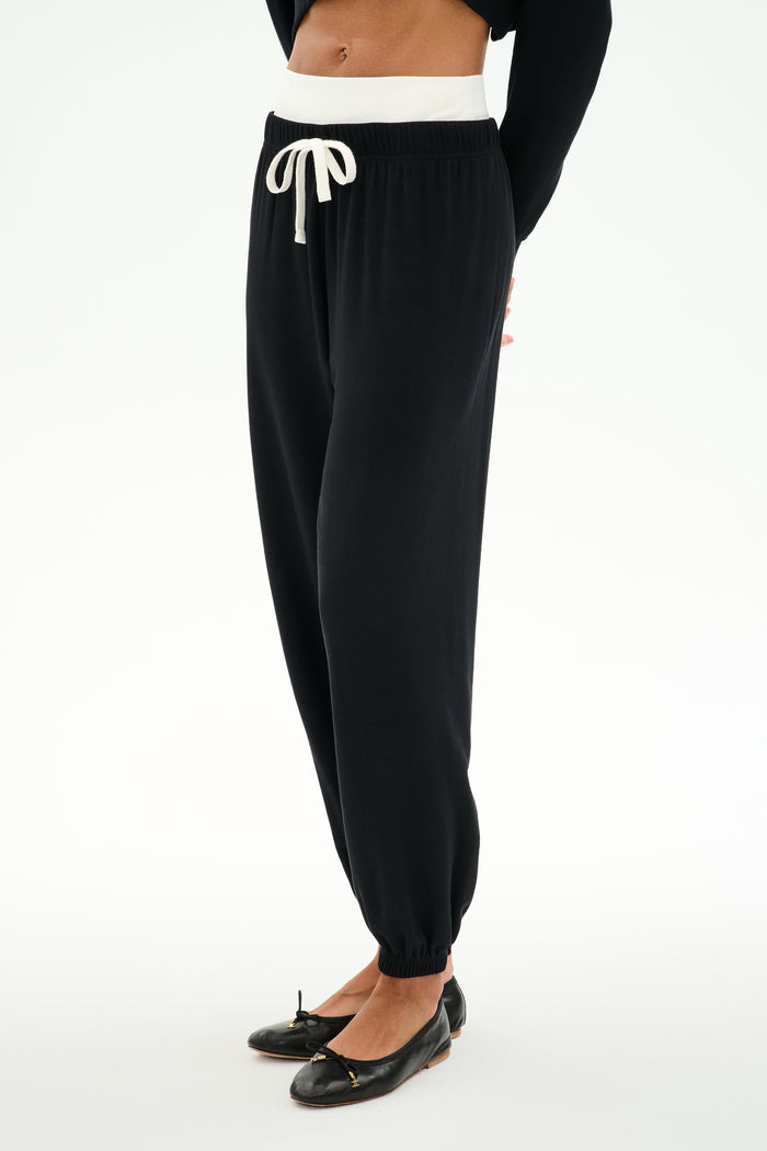A person wears SPLITS59 Andie Dual Waist Sweatpant in Black/White, featuring a white drawstring and waistband, soft modal blend fabric, paired with a black long-sleeve top and ballet flats, standing against a white background.