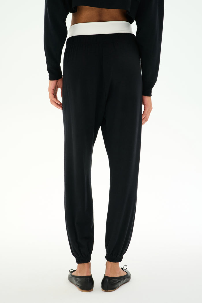Model wears SPLITS59 Andie Dual Waist Sweatpant - Black/White, featuring a soft modal blend with a contrasting white waistband, paired with black flat shoes, shown from the back against a plain white background.
