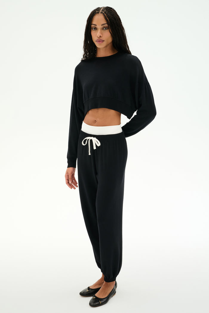 A woman wears a black long-sleeve crop top, the SPLITS59 Andie Dual Waist Sweatpant in Black/White, and black ballet flats, standing against a plain white background.