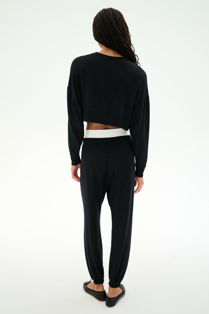 A person with long braided hair faces away, wearing a black modal blend long-sleeve top, black flat shoes, and SPLITS59’s Andie Dual Waist Sweatpant in Black/White against a plain white background.
