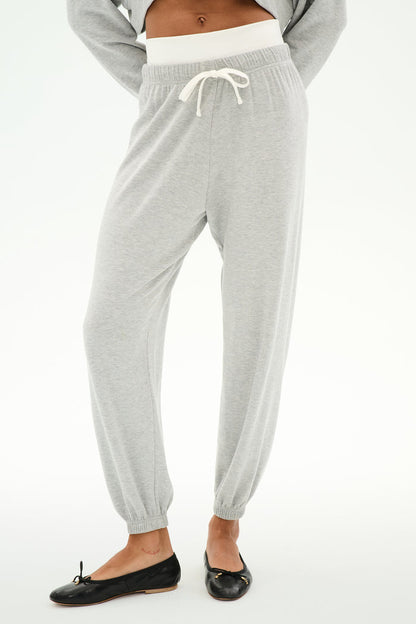 A person wears the SPLITS59 Andie Dual Waist Sweatpant in Heather Grey/White featuring a visible white waistband and white drawstring, paired with black ballet flats, standing against a plain background.