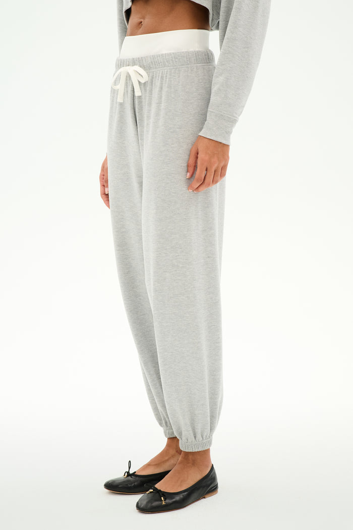 A person wears the SPLITS59 Andie Dual Waist Sweatpant in Heather Grey/White, featuring a white drawstring, paired with a gray top and black ballet flats, standing against a white background.