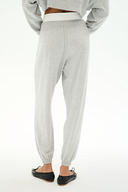 A person wears SPLITS59's Andie Dual Waist Sweatpant in Heather Grey/White with an elastic waist and cuffs, paired with black flats and a cropped gray long-sleeve top, standing on a white background.